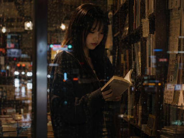 Annetta Hatchcock, met donker haar, in een pluizige grijze trui, leest een boek in een slecht verlichte boekwinkel, staat bij de boekenkasten, kijkt naar beneden en houdt het boek met beide handen vast..