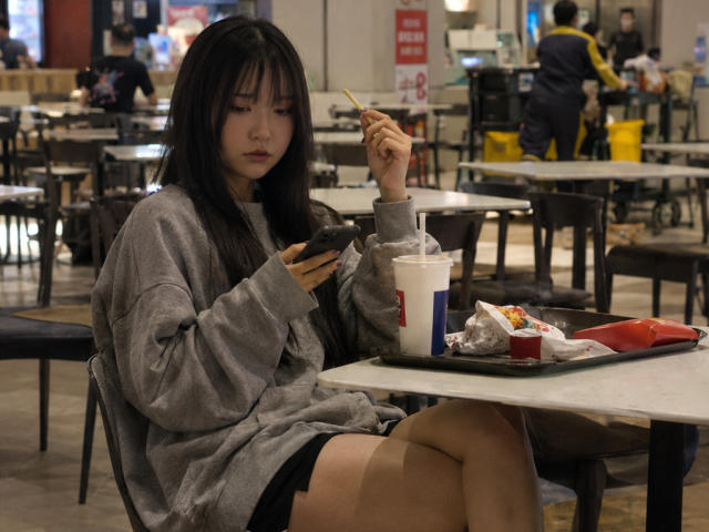 Annetta Hatchcock mit schwarzen Haaren, die einen grauen Pullover und schwarze Shorts trägt, sitzt an einem Tisch, isst und schreibt Nachrichten, hält einen Strohhalm in ihrem Getränk und hat einen Mülleimer neben sich..