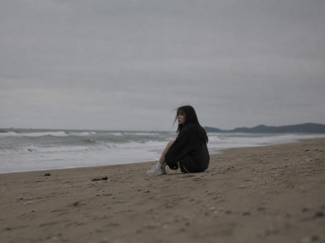 Annetta Hatchcock está sentada em uma praia arenosa, vestindo uma camisa preta e meias brancas, com seu longo cabelo castanho sendo soprado pelo vento, olhando para o oceano..