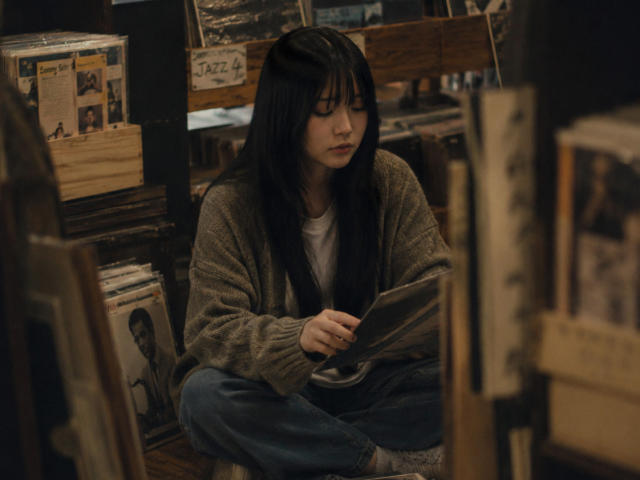Annetta Hatchcock, avec ses longs cheveux noirs, portant un cardigan marron et un jean bleu, est assise en tailleur, lisant un magazine dans une librairie mal éclairée et encombrée..