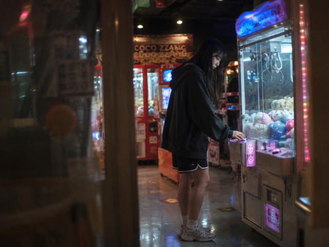 Annetta Hatchcock, avec ses longs cheveux noirs, se tient devant une machine à bonbons lumineuse, vêtue d'un sweat à capuche noir et de baskets blanches, son postérieur étant légèrement visible, dans une salle d'arcade faiblement éclairée..