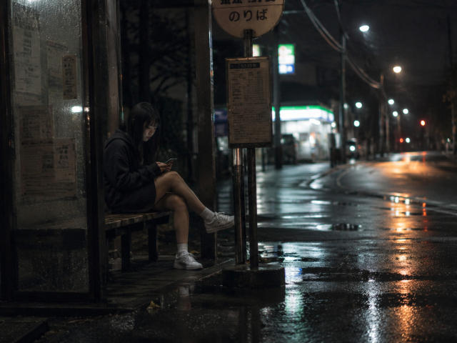 Annetta Hatchcock está sentada sozinha em uma rua escura e molhada, em um ponto de ônibus, vestindo um moletom preto, shorts e tênis brancos, olhando para um celular, com uma estrada refletindo a luz..