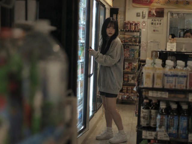 Annetta Hatchcock se tient dans un magasin faiblement éclairé, vêtue d'un sweat à capuche gris, de shorts noirs, de chaussettes blanches et de baskets blanches, et regarde un présentoir réfrigéré. Ses fesses sont visibles..