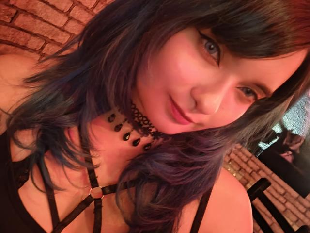 NaommyCarter with blue-purple hair, black choker, and strappy black top showing tits, smiling softly at the camera, in a warm, dimly-lit room.