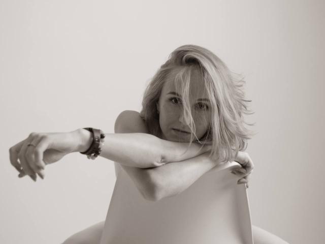Christal Kowalske, met rommelig blond haar, armen over haar blote borst gekruist, kijkt naar de camera, draagt een armbandje om haar linkerpols, zit op een witte stoel, op een zwart-wit foto..