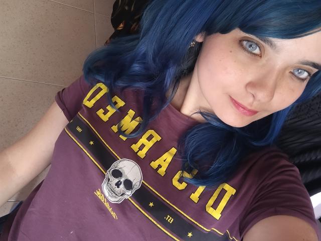 NaommyCarter with blue hair, blue eyes, and light skin, wearing a purple "Ozarka" t-shirt with a skull graphic, taking a selfie. Boobs not visible, pussy not visible.