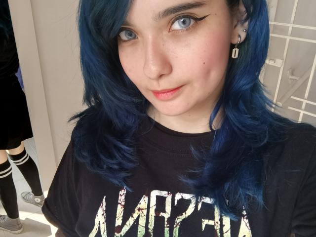 NaommyCarter with blue hair, black shirt, and nose ring, wearing black and white striped socks, has a cute smile, and subtle makeup with black eyeliner.