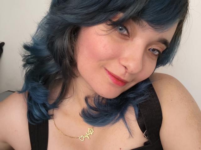 NaommyCarter with blue wavy hair, fair skin, pink cheeks, and red lipstick, wearing a black top and gold necklace, smiling softly at the camera.