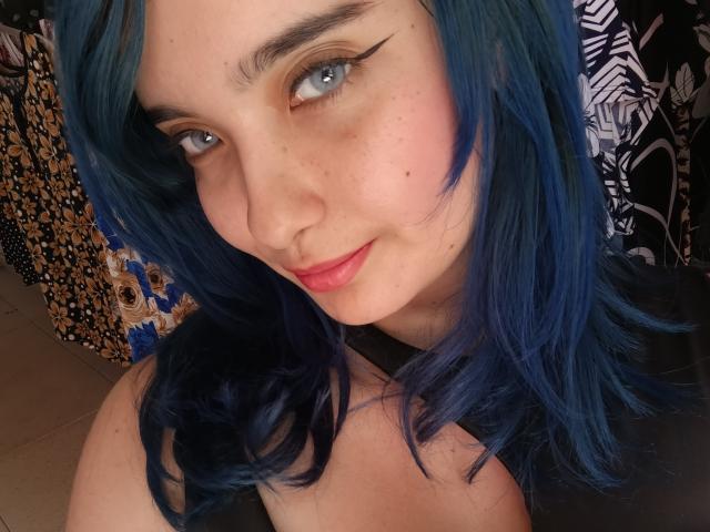 NaommyCarter has blue hair, light skin, and blue eyes with black eyeliner, a nose ring, and pink lips. She's smiling slightly, showing freckles on her face.