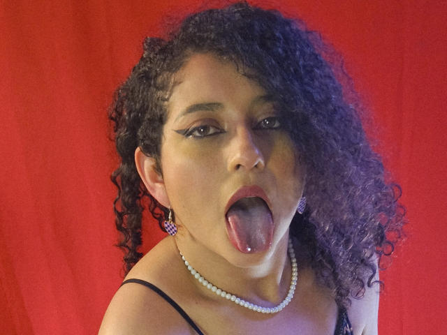 AstraVoss, avec ses cheveux noirs bouclés, sa peau brune et son collier de perles, tirant la langue avec un piercing à la langue, portant un eye-liner noir et des boucles d'oreilles roses, sur un fond rouge..