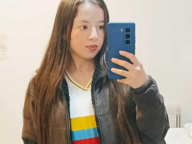 VioletaNova met lang bruin haar, die een zwarte jas draagt over een wit shirt met gele, blauwe en rode strepen, maakt een spiegelzelfportret met een blauwe telefoon..