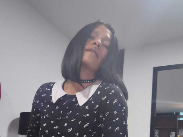 AmelieBlake with shoulder-length black hair, nose ring, black choker, and black dress with white collar and small white patterns, slightly tilting head back, smiling.