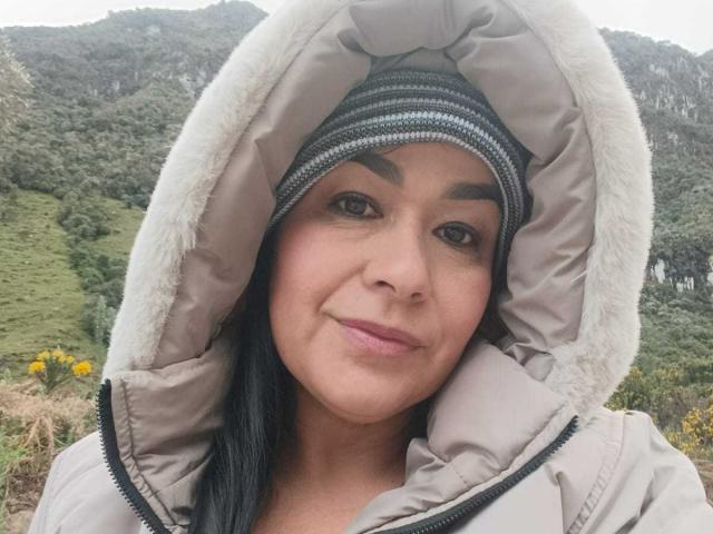 TamaraFoxy with dark eyebrows, black hair, and pink lips, wearing a beige puffy jacket with a furry hood, and a striped knit beanie, smiling softly.