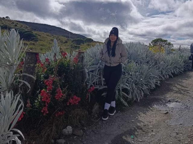 TamaraFoxy stands in a gravel path, wearing a beige jacket, black beanie, dark pants, and white socks with black sneakers. She's surrounded by gray agave and red flowers.