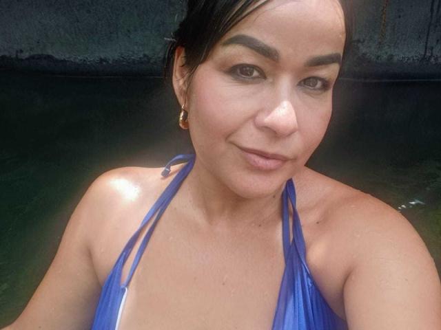 TamaraFoxy with wet, brown skin, small tits in a blue halter top, dark hair, eyebrows, and a subtle smile, standing in green water, wearing gold earrings.