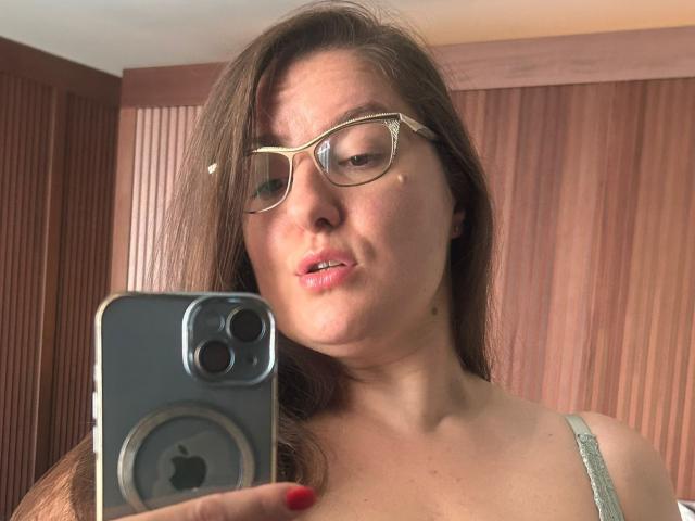 BadAngel with glasses, holding an iPhone, taking a selfie, showing her face, slightly parted lips, red nails, and a green strap, brown hair, and a wooden background.