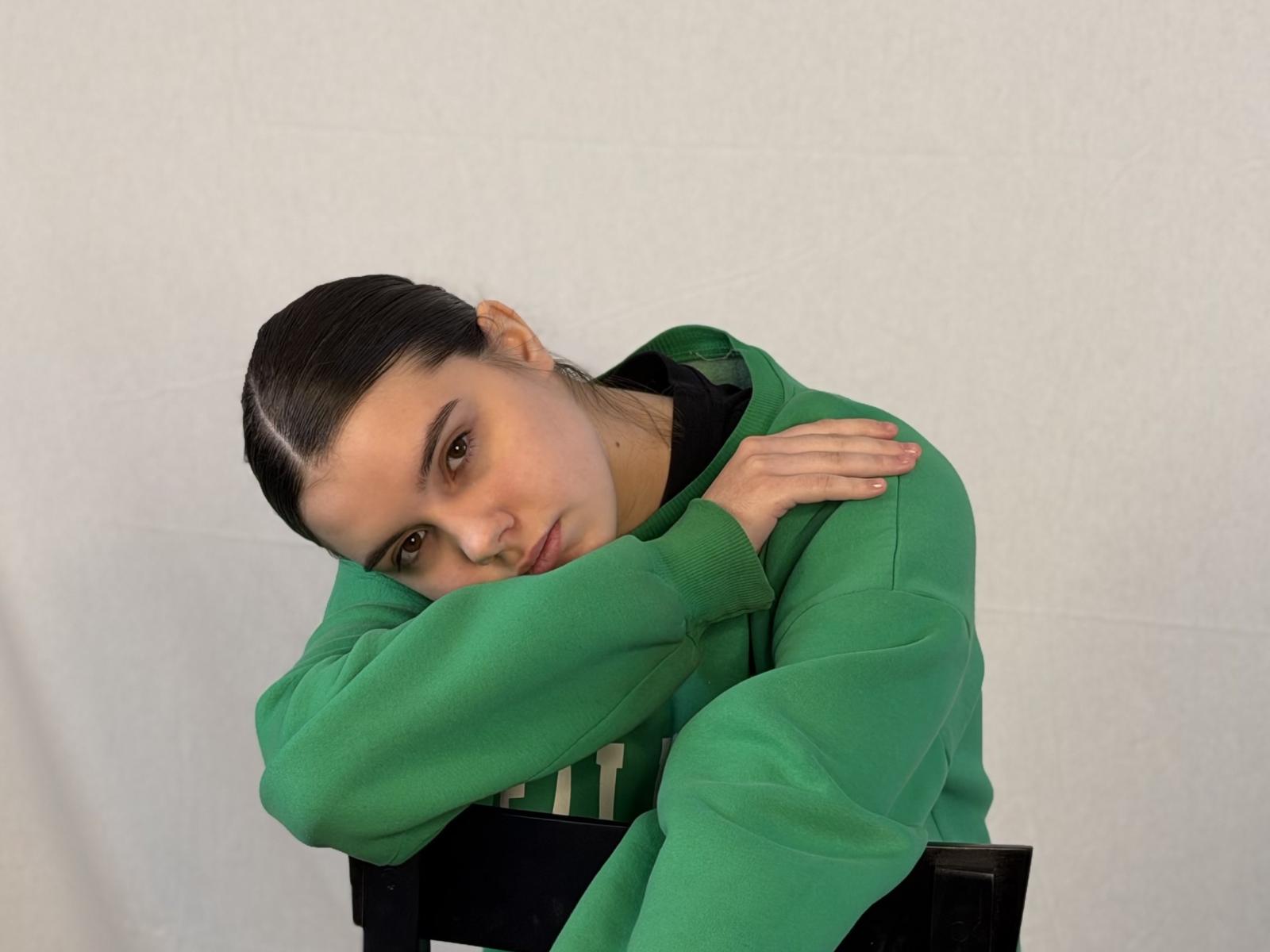 Telma Lavey, com cabelo preto penteado para trás, moletom verde e blusa preta, braços cruzados, cabeça apoiada na mão, unhas verdes, expressão triste, fundo de parede branca..