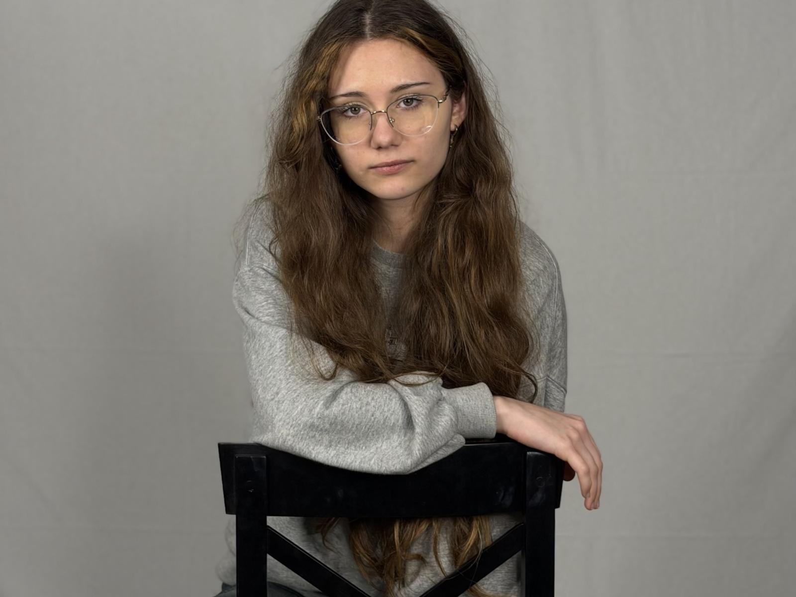 Ellyn Koscinski, con lunghi capelli castani, occhiali, un maglione grigio e seduta su una sedia nera, con le braccia appoggiate sulla sedia, che guarda leggermente di lato..