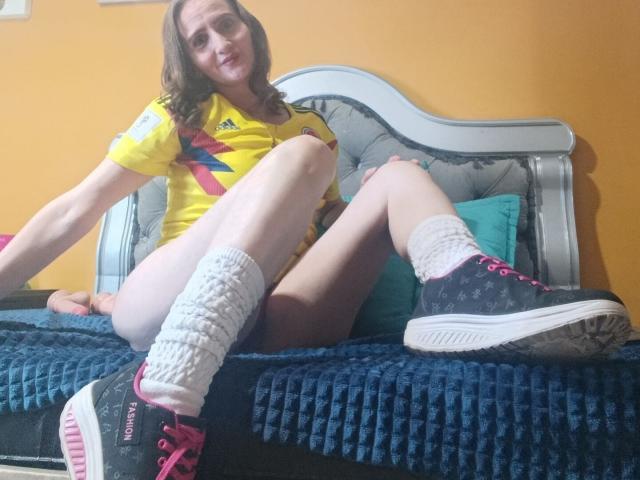 RubiAnderson with pale skin, brown hair, wearing yellow soccer shirt, white leg warmers, black sneakers with pink laces, sitting on blue bed, legs spread, showing her bare pussy.
