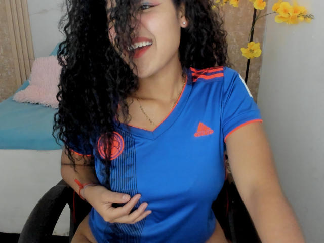 AriaVega with curly black hair, smiling, wearing a blue shirt with red trim, covering her tits with one hand, sitting on a black chair, yellow flowers in background.