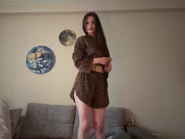BreanaAdina stands in a leopard print dress, arms crossed, showing her legs, with long brown hair, in a room with a gray couch and two wall decorations.