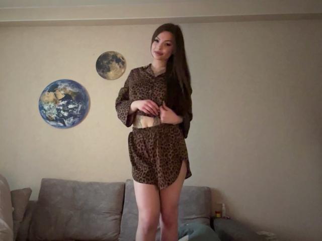 BreanaAdina with long brown hair, wearing a leopard print dress, standing in a room, hands on her belly, smiling, showing her ass, in front of a gray couch.