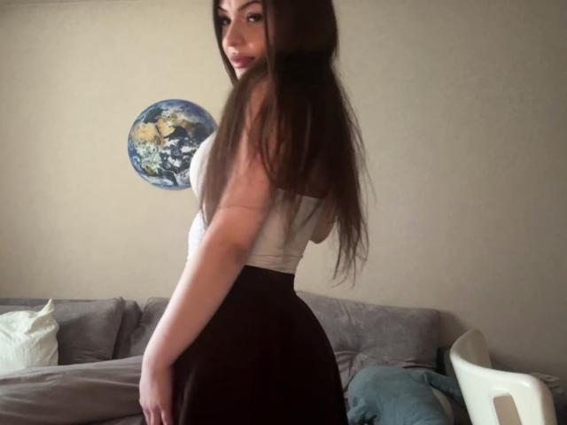 BreanaAdina with long brown hair, white tank top, and black skirt, standing sideways, showing her ass, looking back with a slight smile, in a dimly lit room.