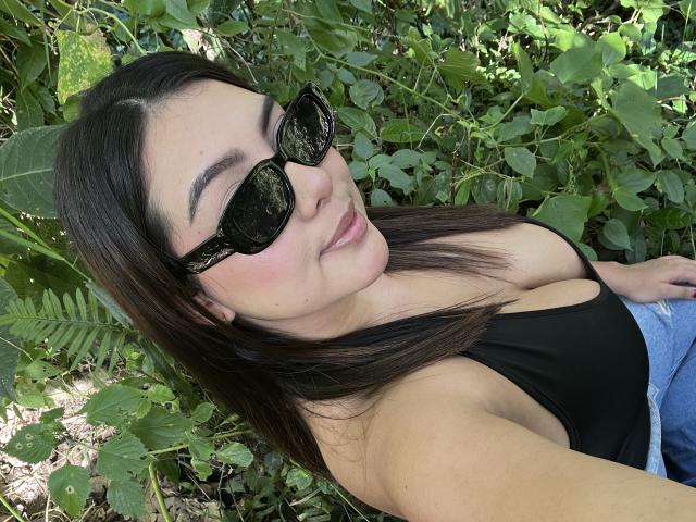 AlannaL com óculos de sol pretos, cabelo castanho liso, top preto, mostrando os seios, deitada em folhagem verde, sorrindo levemente, jeans azuis, pele clara, relaxada, selfie casual..