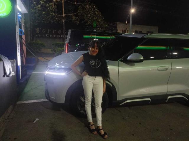 DianaLane with small tits, wearing a black sleeveless top, white skinny pants, and black heels, leans against a white SUV at night, hands on hips, smiling.