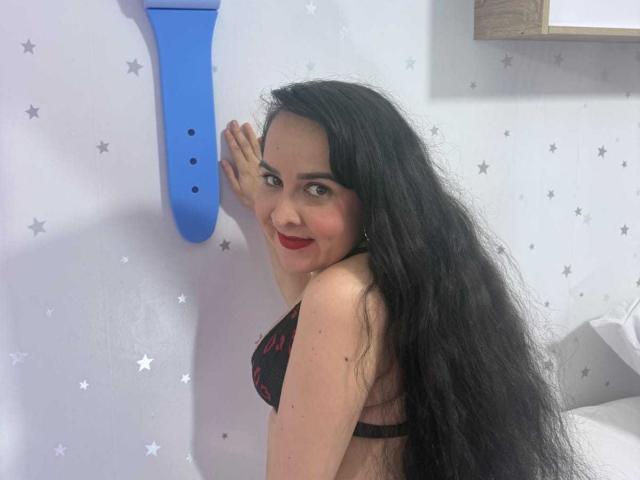 Diana Lane met lang zwart haar, rode lippenstift, een zwarte kanten bh, lachend voor een blauwe muur, waarbij haar borsten zichtbaar zijn, in een kamer met een wit sterrenpatroon..