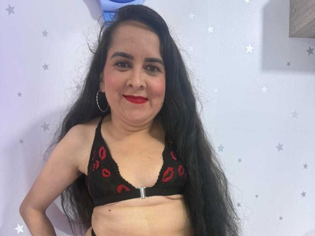 DianaLane with long black hair, red lipstick, black lace bra with red kiss prints, small tits, slight belly, smiling, hoop earrings, white wall with stars.