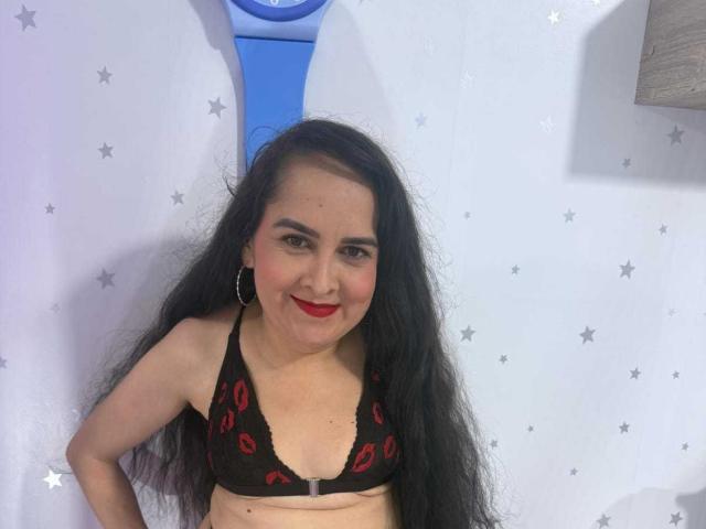 Diana Lane met lang zwart haar, die een zwarte kanten BH draagt met rode lippenstiftsmotieven, lacht naar de camera, toont haar kleine borsten, rode lippenstift en oorringen..