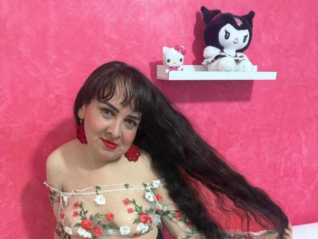 Diana Lane met lang zwart haar, rode oorbellen en een doorschijnende top met bloemenborduurwerk, waarbij haar borsten zichtbaar zijn, lachend, voor een roze muur met Hello Kitty en My Melody op een plank..