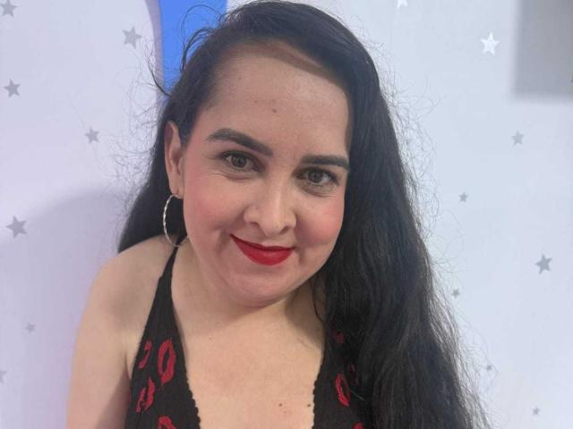 Diana Lane met lang zwart haar, rode lippenstift, een zwart topje met halterhals en rode patronen, oorringen en een schattige glimlach, waarbij haar borsten lichtjes zichtbaar zijn op de foto..