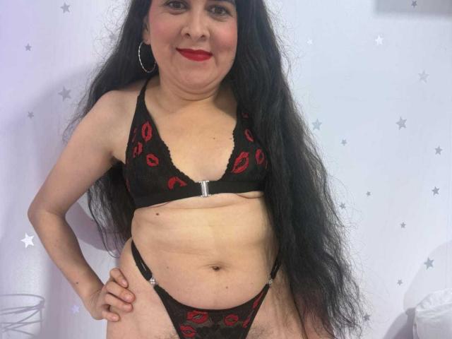 Diana Lane, met lang zwart haar en rode lippenstift, draagt zwart kant lingerie met rode lippenstiftmotieven, staat met haar handen op haar heupen, toont haar borsten en haar schaamdelen zijn licht zichtbaar..