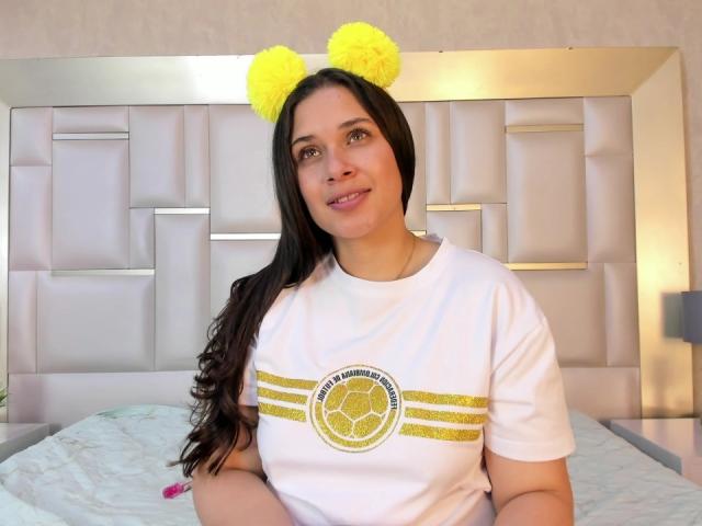 EmmyRossi has yellow fluffy ears on her head, long black hair, and a white shirt with gold stripes and a soccer logo, sitting on a bed, smiling.