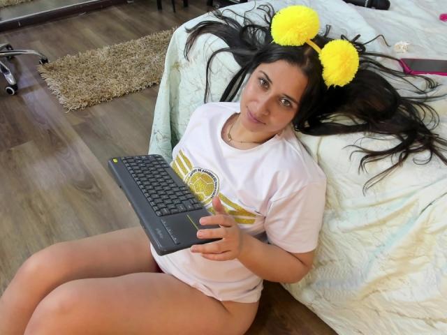 EmmyRossi con orecchie a forma di pompon gialle, seduta su un pavimento di legno, indossa una maglietta bianca e non ha pantaloni, tiene un portatile nero, ha capelli lunghi e scuri, un sedere piccolo, sta sorridendo, camera da letto..