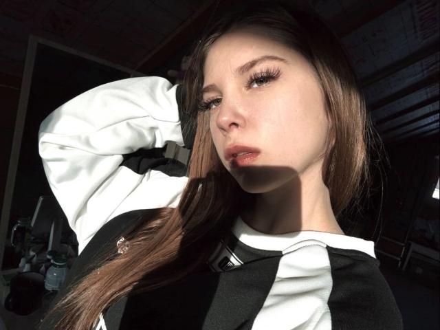 Danae Egger, met lang bruin haar, draagt een zwart-witte top en heeft haar arm omhoog. Het zonlicht accentueert haar gezicht, waardoor haar lange wimpers en licht open roze lippen zichtbaar zijn..