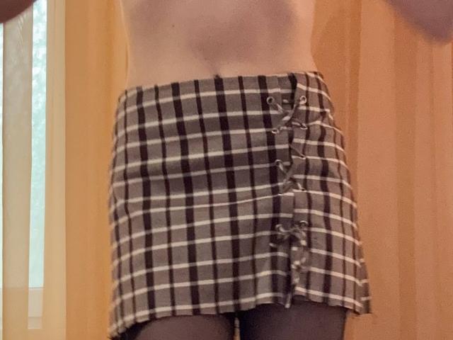 La falda escocesa de MiyaLia, con cuadros negros, blancos y grises, cordones en la parte delantera y medias negras, que muestra su abdomen plano y parte de su trasero desnudo..