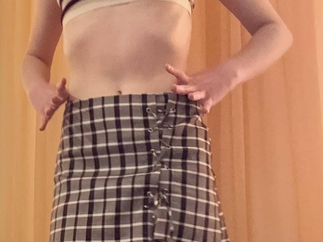 MiyaLia con la pancia piatta, mani su una gonna a quadri, abbottonata davanti, che lascia intravedere un accenno del suo sedere, indossa un reggiseno beige, su uno sfondo di legno..