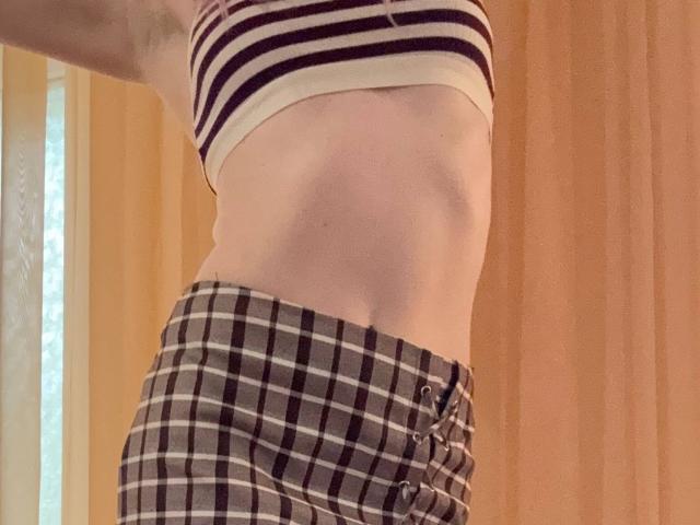 La figura delgada y pálida de MiyaLia, con un abdomen plano y abdominales apenas marcados, viste una camiseta corta a rayas y pantalones a cuadros, mostrando sus glúteos pequeños y planos y pantalones abotonados..