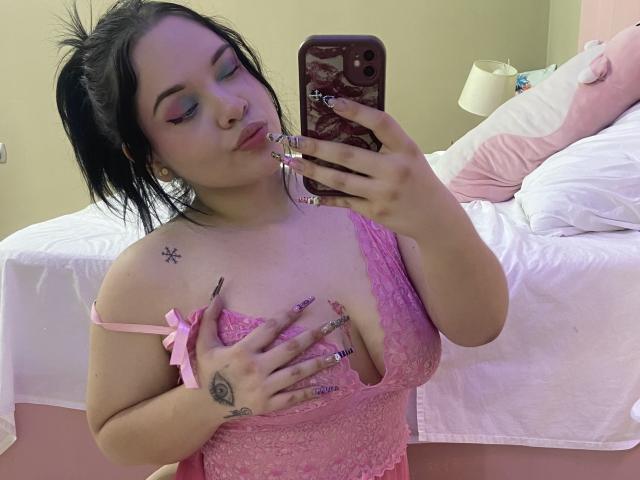 AlayaWillow with black hair in a ponytail, pink lace lingerie, holding phone, covering her tits with her hand, pink nails, tattoo on shoulder, tattoo on hand, in bedroom.