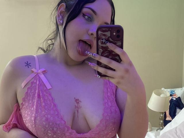 AlayaWillow with black hair, pink lace bra, tongue out, holding phone with purple case, pink ribbon, tattoo on chest, and tattoo on shoulder, showing her tits and nipple.