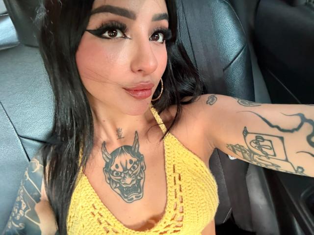 ScarletFeline mit schwarzen Haaren, einem gelben, gehäkelten Top und Tattoos, darunter ein Wolfsschädel auf der Brust, sitzt in einem Auto, lächelt und trägt goldene Creolen, wobei sie ihre Brüste zur Schau stellt..