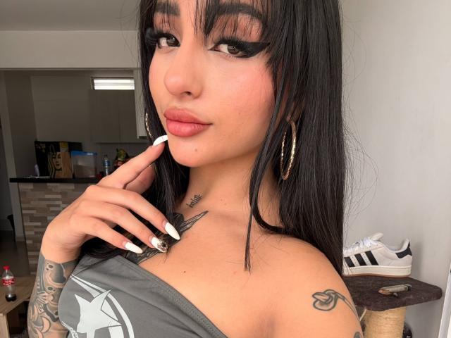 ScarletFeline, com cabelos pretos longos, cílios grossos, batom rosa, brincos de argola dourados, tatuagens no pescoço e no braço, tocando o queixo, vestindo uma blusa cinza com um ombro descoberto, unhas brancas e tênis brancos..