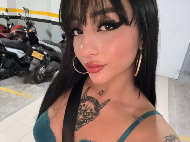 ScarletFeline tem cabelo escuro, delineador marcante e batom rosa. Ela está usando um sutiã verde, brincos de argola e uma tatuagem de caveira no peito. Seus seios estão visíveis..