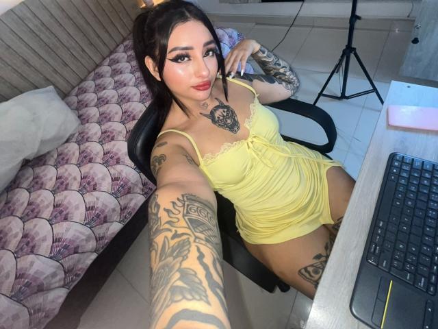 ScarletFeline, com tatuagens nos braços e no peito, vestindo um vestido amarelo, sentada em uma cadeira, tirando uma selfie, mostrando seus seios, nádegas e vulva, em um quarto com uma mesa..