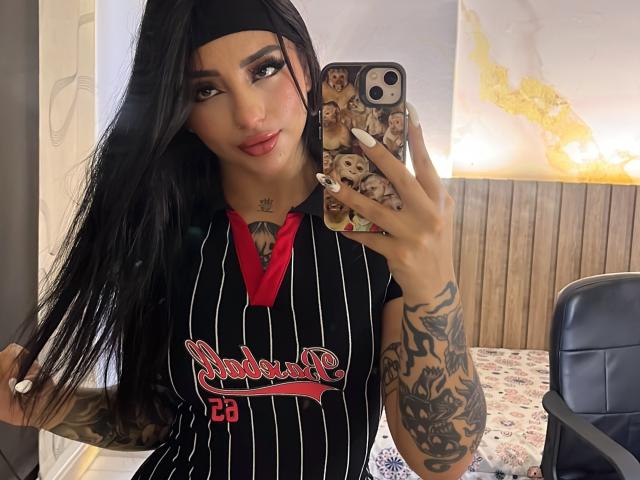 ScarletFeline, con lunghi capelli neri, indossa una camicia nera a righe e tiene in mano un telefono con una custodia a forma di cane. Ha tatuaggi sul braccio e sul petto e si sta guardando allo specchio..