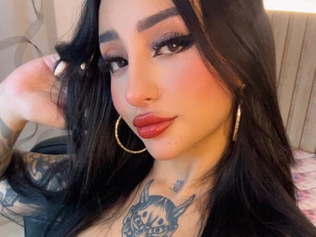 ScarletFeline tem cabelo escuro, sobrancelhas marcadas, batom vermelho, brincos de argola, tatuagens e um piercing no nariz. Sua maquiagem é dramática, com cílios longos e um olhar levemente esfumado..