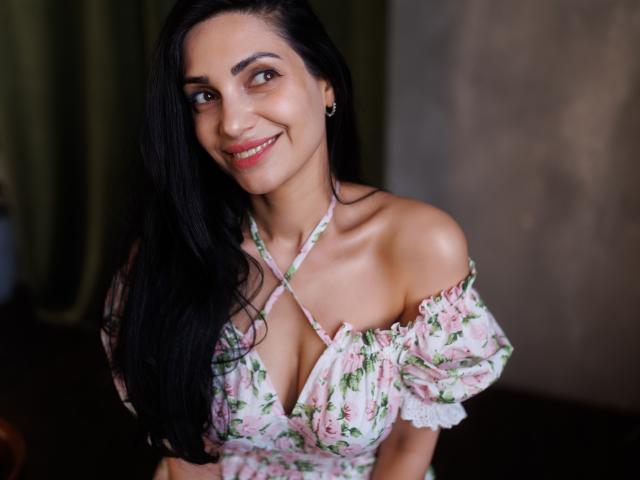Beatrice, capelli lunghi e neri, sorridente, indossa una blusa floreale con una spalla scoperta che mette in mostra il seno, rossetto rosa e piccoli orecchini, in un ambiente interno con una luce soffusa..